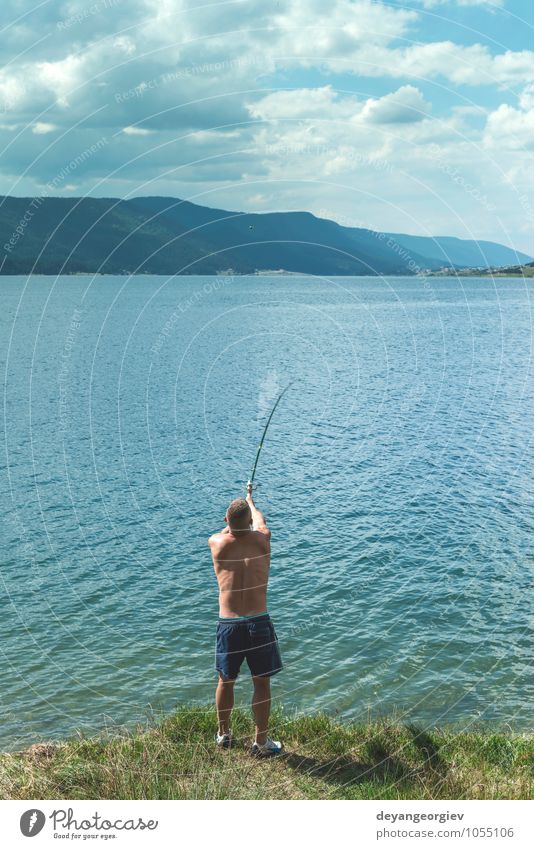 A young man with a fishing rod stands on a bank next to a waterfall, next  to him is a young man who is reaching out his hand to a swimming young