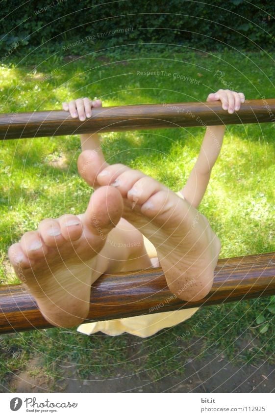 Small child hangs clamped to a wooden bar, outside in the nature. Girl is doing gymnastics on a bar, holding on with her hands and stretching her bare feet upwards. Funny feet, appear oversized and bizarre through perspective.