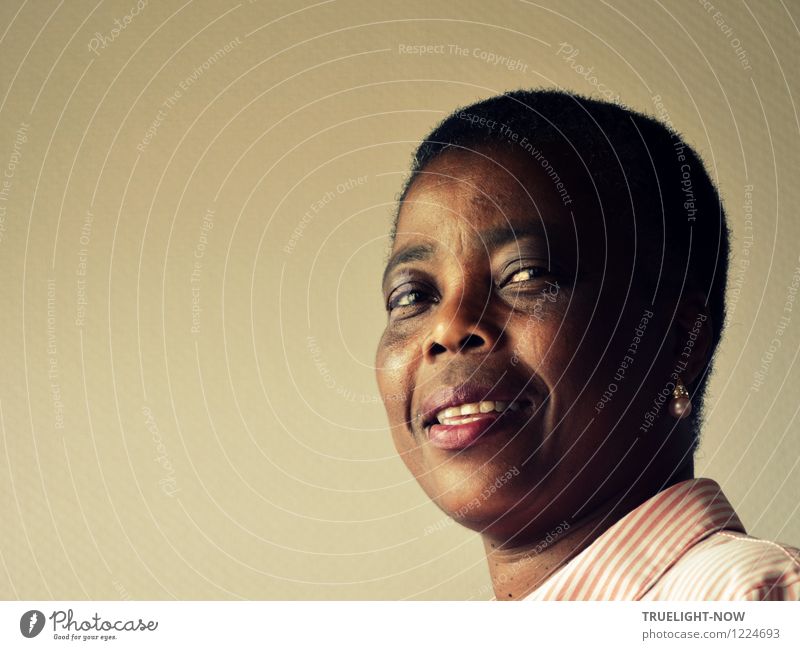 Beautiful, strong African woman in half profile with short hairstyle, pearl on ear and white and pink striped blouse looks smiling into the camera with slightly opened mouth