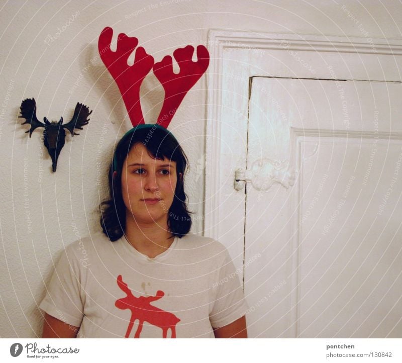Disguise. Young woman wearing a toy antlers stands in front of a fake moose antlers and wears a t-shirt with a moose motif. Repeat. humor, bizarre