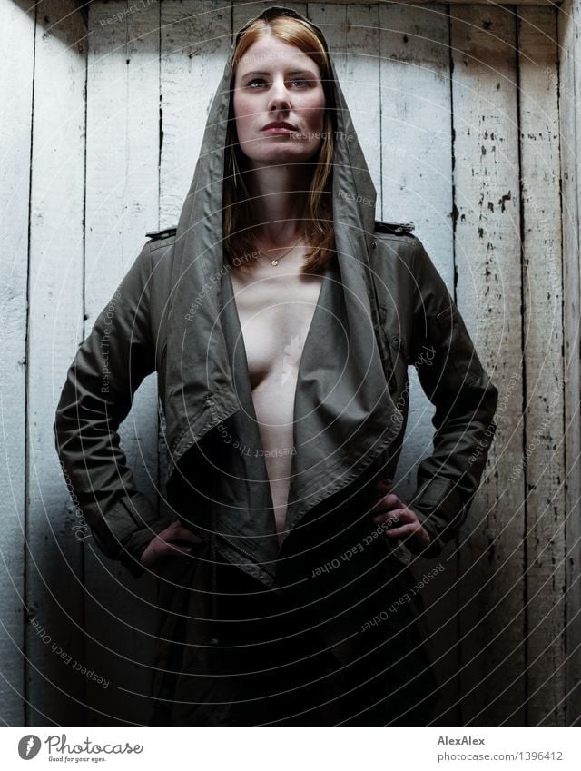 A tall, red-haired woman stands in front of an old wooden wall in an attic, dressed only in a coat and illuminated by the skylight Wooden wall Attic Young woman