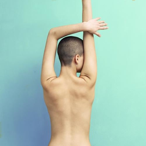 young woman with very short hair stands naked in front of light turquoise wall in pose with half raised arms Athletic Fitness Harmonious Well-being Meditation