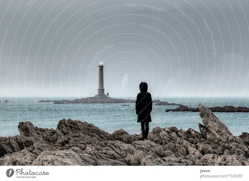Woman in coat with hood observes distant lighthouse in sea Lighthouse Rock coast person rocky curt harsh somber Hope Longing Observe Ocean Water Horizon