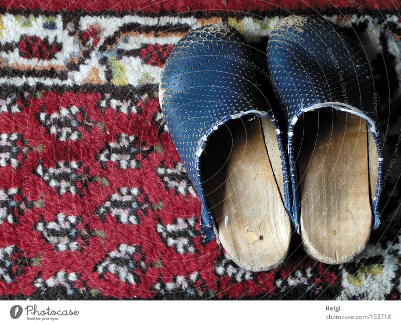 a pair of worn-out wooden shoes standing on an old patterned carpet Footwear Wood Leather Old Broken Shabby Worn out Carpet Pattern Red Blue White Clothing