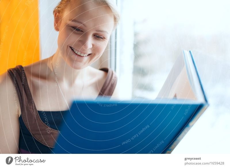 Beautiful young smiling woman reading a book with blue cover while sitting at a window Lifestyle Happy Relaxation Reading House (Residential Structure)