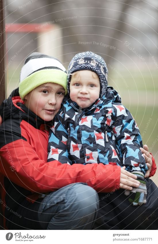 Two adorable young brothers outdoors in winter wrapped up warmly against the chill weather with the older boy cuddling his toddler sibling on his lap Joy Garden