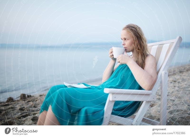 Wide shot of young girl enjoying a cup of tea at the seaside sitting relaxing on a deckchair with a blissful expression overlooking a tropical beach Beverage