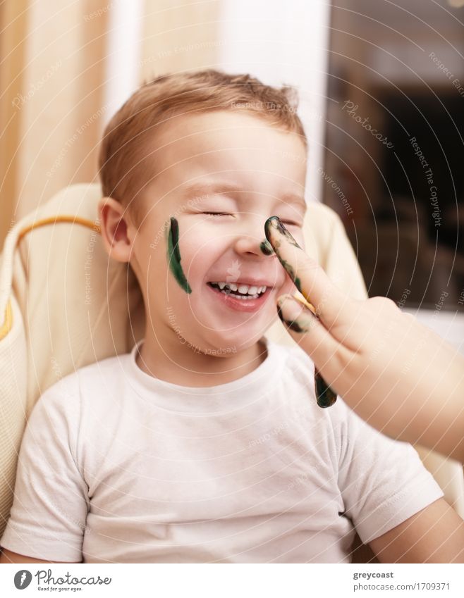 Little boy laughing as his mother paints his face streaking his cheek and dabbing his nose with black face paint Joy Happy Leisure and hobbies Playing Child