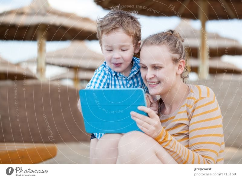 Happy mother and her small son at a beach resort playing on a tablet computer together under beach umbrellas at the seaside Joy Playing Beach Entertainment