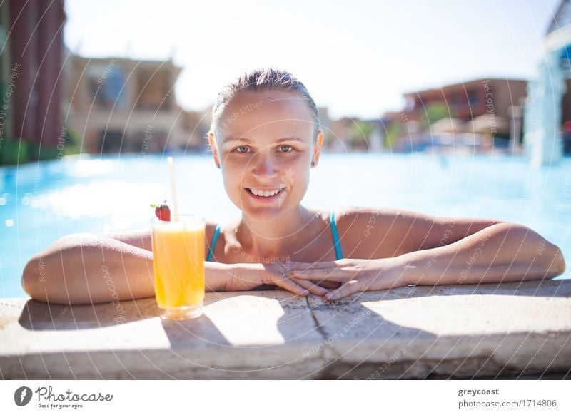 Smiling young woman enjoying a refreshing orange drink or cocktail in the pool resting her arms on the pool surround smiling at the camera Beverage Drinking