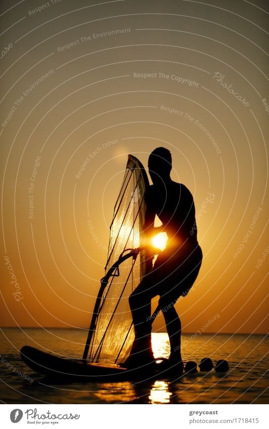 Man sailboarding at sunset silhouetted against the bright orb of the sun in a colorful orange sky on a calm ocean Relaxation Vacation & Travel Freedom Summer