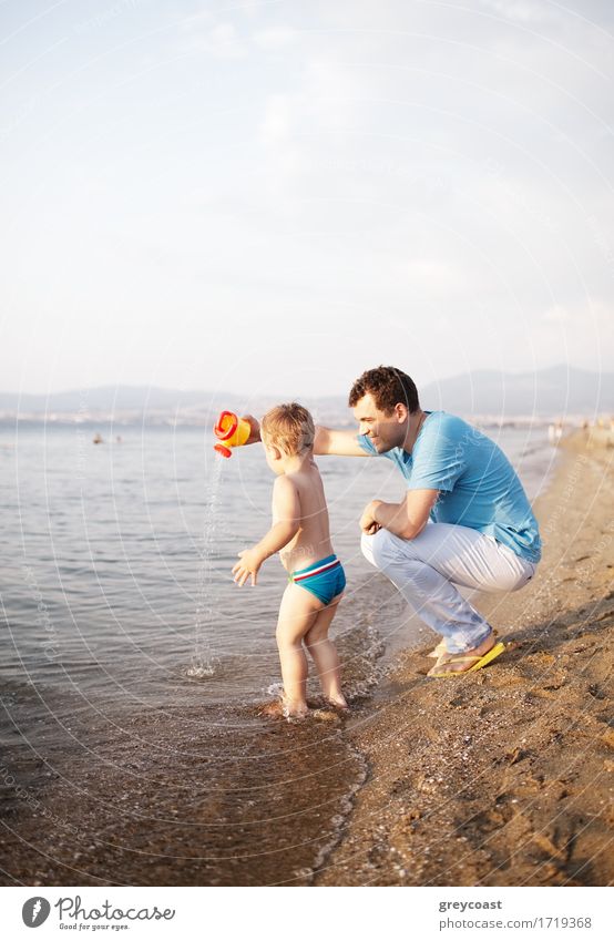 Young father playing with his son at the beach crouching down a the edge of the water as the toddler happily paddles in the shallow water playing with a watering can