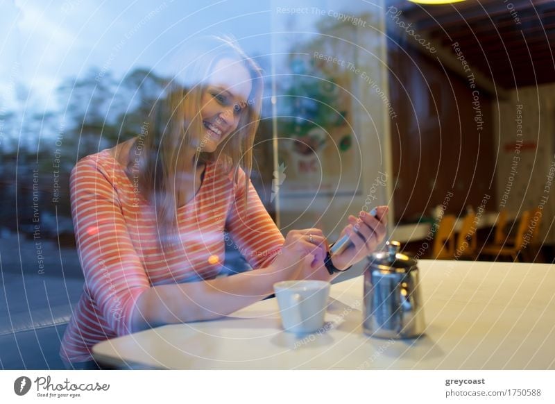 View through the glass window of a young woman using a mobile phone in a cafeteria smiling as she reads a text message Hot drink Coffee Tea Happy Restaurant