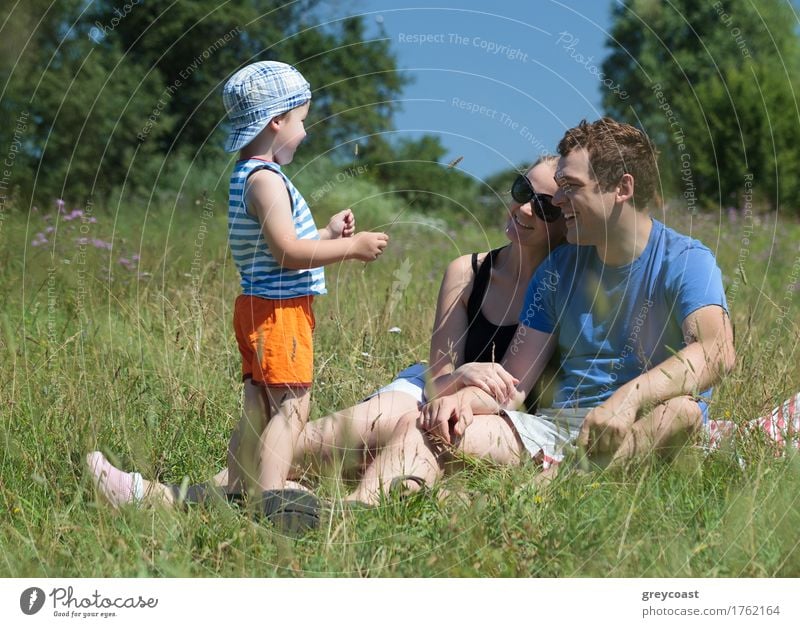 Young parents and son on the meadow on a hot summer day. Happy couple sitting on the grass and looking at the kid. Outdoor family activity Joy Relaxation