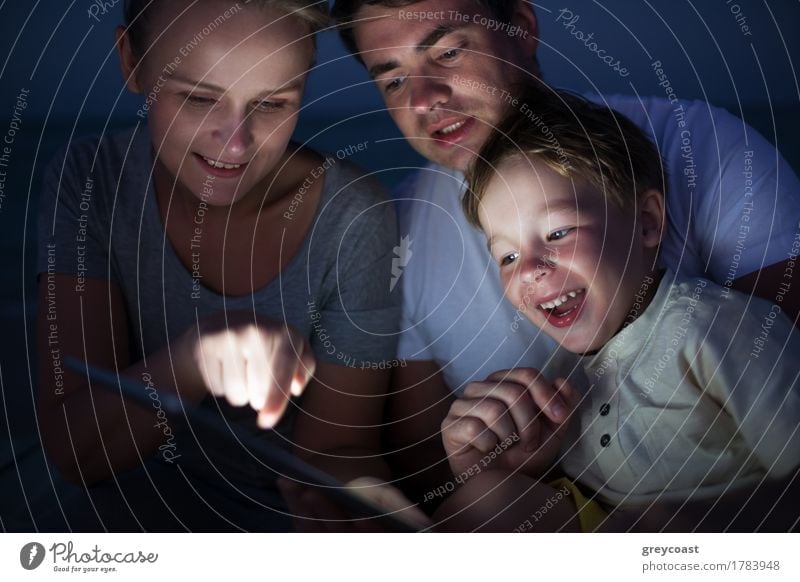 Happy family of three with pad late in the evening. Laughing child looking at screen where mother pointing at something Joy Leisure and hobbies Playing
