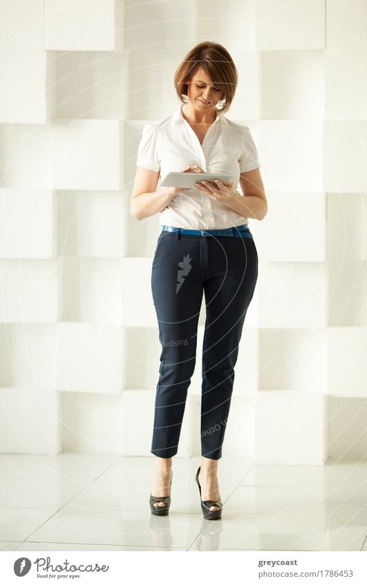Smiling businesswoman standing against white wall while looking at tablet in her hand Lifestyle Elegant Style Office Business Adults Hand Elements Places Shirt