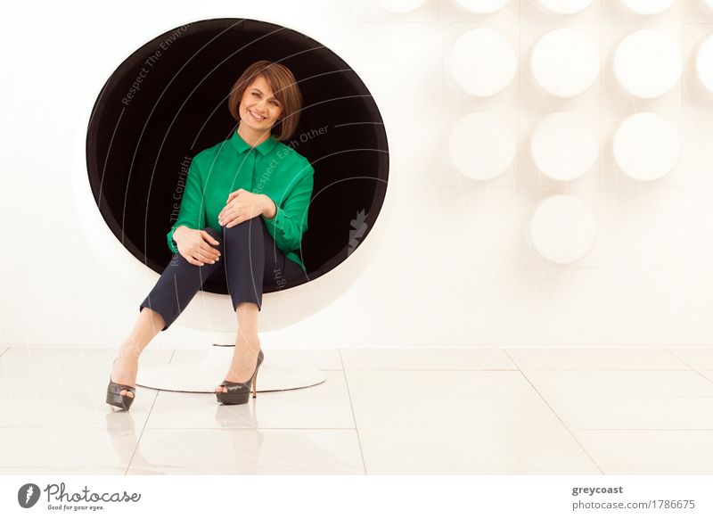 Attractive adult woman sitting on spherical chair with hands on knees. Modern white wall on background Lifestyle Elegant Style Chair Office Human being Woman