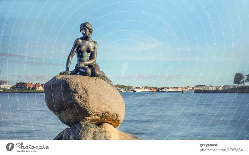 Front view of Little Mermaid statue on large boulders in Denmark with harbor under blue sky in the background Vacation & Travel Tourism Ocean Rock Harbour