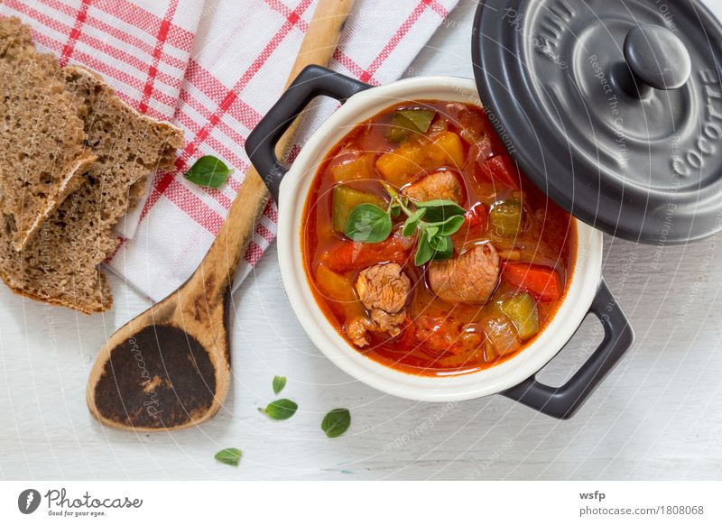 Goulash in a black cocotte with marjoram on white wood Bread Soup Stew Wooden spoon Dark Black White gulyas gollash gujash Marjoram Hungarian Eating Pepper