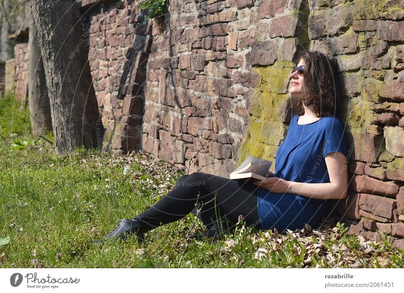 Soaking up the sun - young brunette woman sitting in a park in the grass leaning against a stone wall with a book in her hand and enjoying the sun Human being