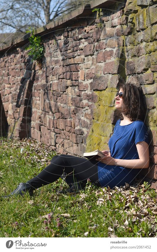 Soaking up the sun - young brunette woman sitting in a park in the grass leaning against a stone wall with a book in her hand and enjoying the sun Well-being