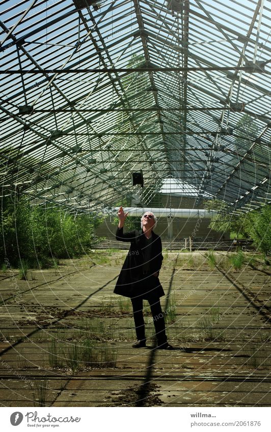 A man in a frock coat casually tosses his top hat into the air Man Frock coat Cylinder Sunglasses Hat Greenhouse White-haired Throw Bushes Full-length