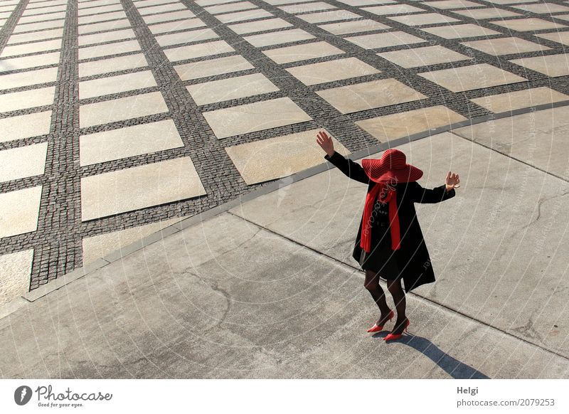 elegantly dressed lady with black coat, red hat, red scarf and red pumps is dancing on a large square with concrete and patterned floor Human being Feminine