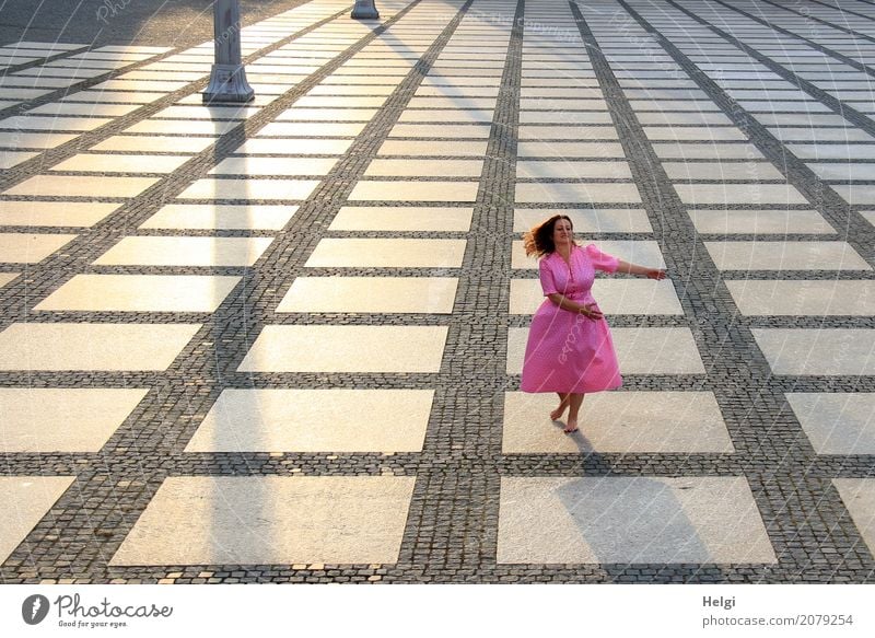 Woman with long brunette waving hair and pink dress dancing barefoot on a large paved square against the light Human being Feminine Adults 1 45 - 60 years Town