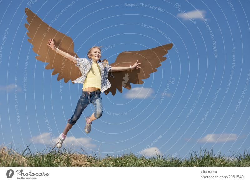 Little girl playing with cardboard toy wings in the park at the day time. Concept of happy game. Child having fun outdoors. Picture made on the background of blue sky.