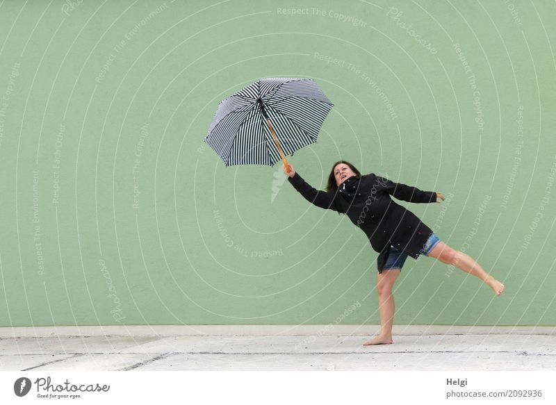Woman with black jacket and short jeans stands barefoot on one leg in front of a light green wall and holds up an umbrella with one arm Human being Feminine