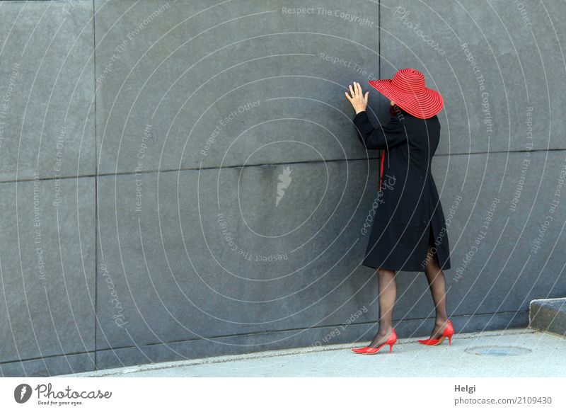 rear view of an elegantly dressed lady with black coat, red hat and red pumps in front of a grey wall Human being Feminine Woman Adults Female senior