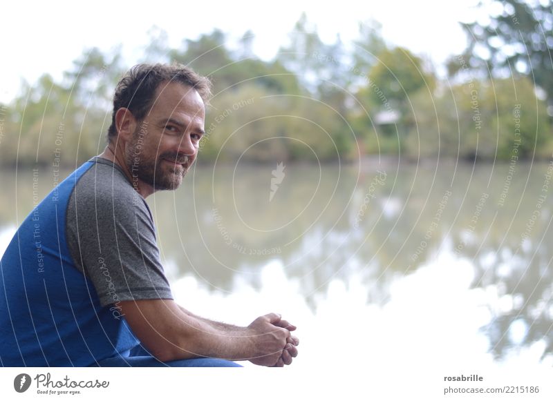 middle-aged dark-haired man with beard squats in a clearing by the pond and looks over his shoulder into the camera Leisure and hobbies Human being Man Adults 1