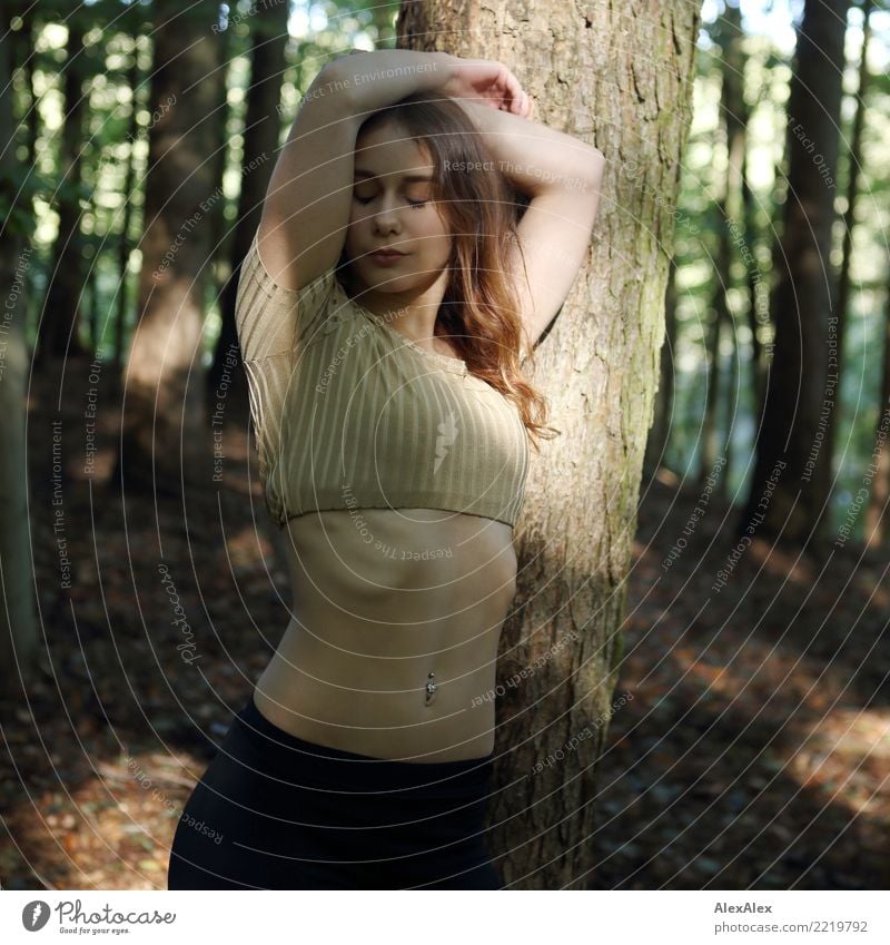 Belly feeling - a young, sporty woman with a belly-free top leans against a tree with her arms folded above her head, closed eyes and a belly button piercing in her muscular, flat belly - light and shadow play in the forest
