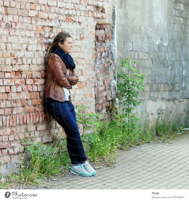 thoughtful woman with dreadlocks, in jeans and brown leather jacket is standing at an old dilapidated house wall Human being Feminine Woman Adults 1