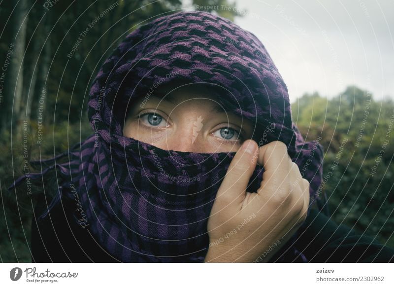 Chica de ojos azules mirando a camara tapandose la cabeza y parte de la cara con un pañuelo aguantado por su mano en la naturaleza. Lifestyle Exotic Face