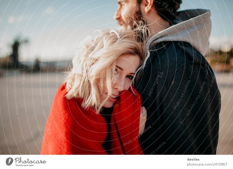 Young blonde woman looking at camera while embraces her boyfriend outdoors. Lifestyle Joy Sun Winter Woman Adults Man Couple Fashion Blonde Smiling Love Embrace