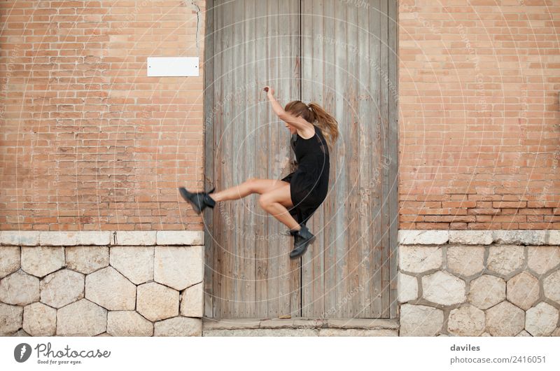 Blonde woman in black dress dancing and jumping in the street, in front of an old house facade. Elegant Style Entertainment Dance Human being Young woman