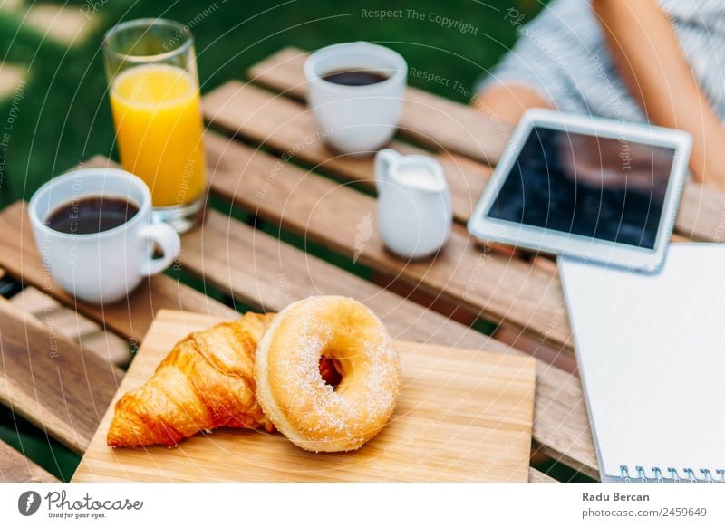 Young And Attractive Woman Having Morning Breakfast In Green Garden With French Croissant, Donuts, Coffee Cup, Orange Juice, Tablet and Notes Book On Wooden Table