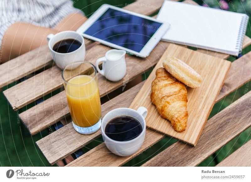 Young And Attractive Woman Having Morning Breakfast In Green Garden With French Croissant, Donuts, Coffee Cup, Orange Juice, Tablet and Notes Book On Wooden Table