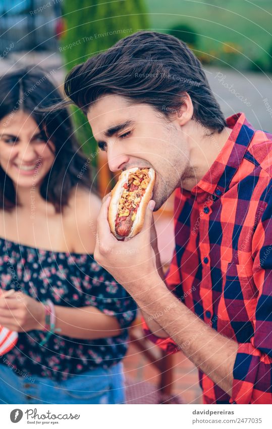 Young man eating hot dog and woman laughing in background Sausage Bread Roll Lunch Fast food Lifestyle Joy Happy Summer Woman Adults Man Friendship Hand Group