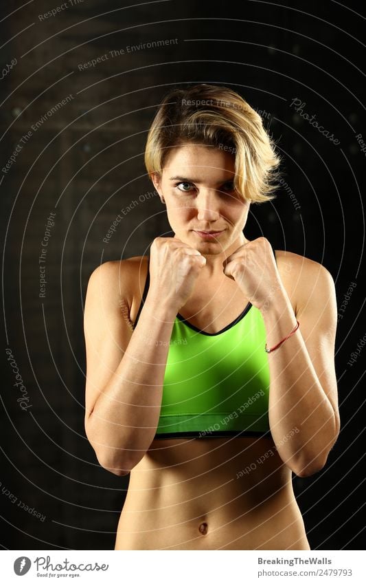 Close up front portrait of one young athletic woman in sportswear in gym over dark background, standing in boxing stance with hands and fists, looking at camera