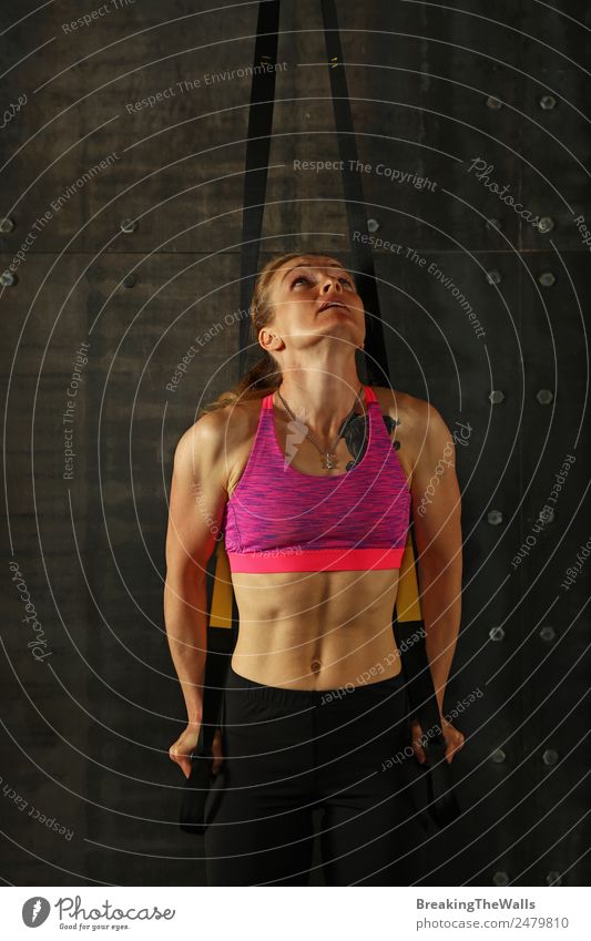 One young middle age athletic woman at crossfit training, exercising with trx suspension fitness straps over dark background, front view, looking up Sports