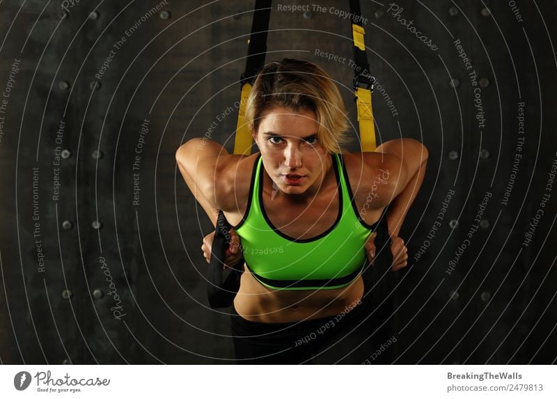 One young athletic woman at crossfit training, exercising with trx suspension fitness straps over dark background, front view, looking at camera Lifestyle