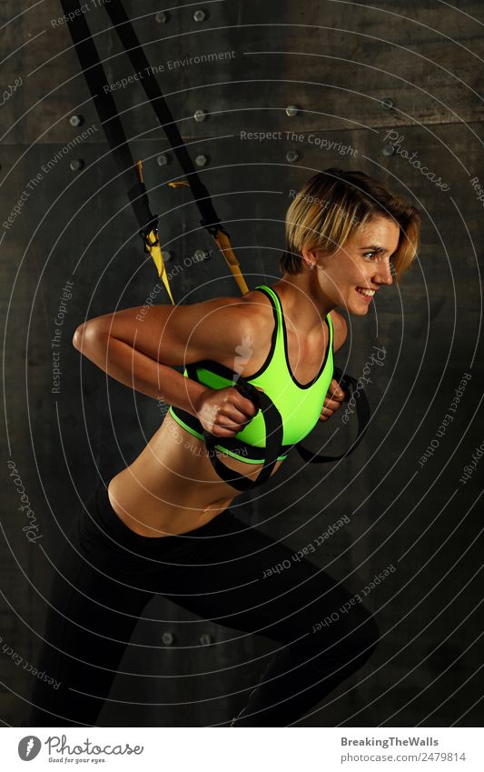 Side profile view portrait of one young athletic woman at crossfit training, exercising with trx suspension fitness straps over dark background, looking away