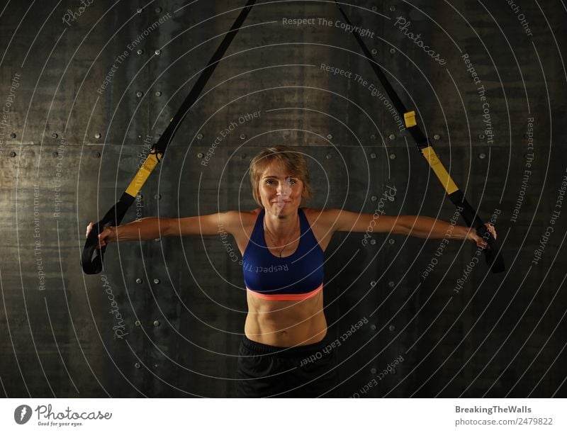One young middle age athletic woman at crossfit training, exercising with trx suspension fitness straps over dark background, front view, looking at camera