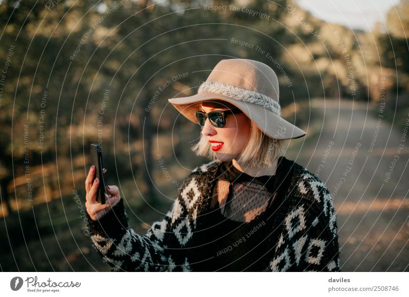 Blonde young woman with hat and sunglasses taking a photo with a mobile phone in the forest at sunset. Lifestyle Elegant Style Relaxation Telephone Cellphone