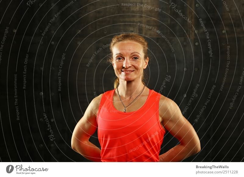 Close up front upper body portrait of one middle age athletic woman in sportswear in gym over dark background, looking at camera and smiling Lifestyle Sports