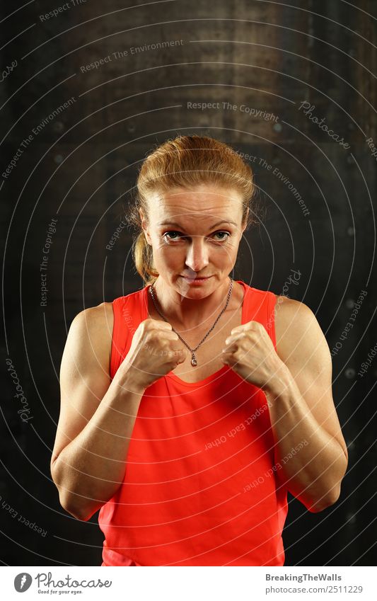Close up front portrait of one young mid adult athletic woman in sportswear in gym over dark background, standing in boxing stance with hands and fists, looking at camera