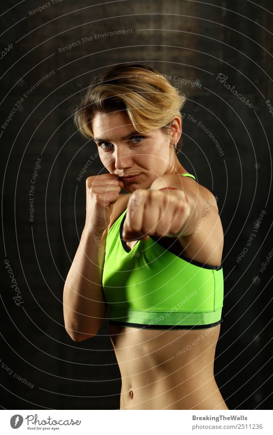 Close up front portrait of one young athletic woman in sportswear in gym over dark background, standing in boxing stance with hands and fists, looking at camera