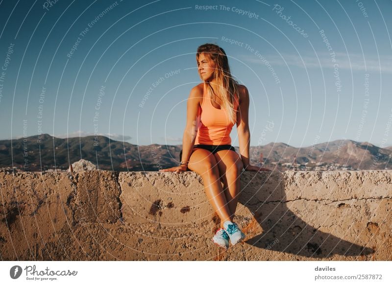 Beautiful woman with sports clothes, sitting on a concrete wall outdoors at sunset. Lifestyle Body Athletic Wellness Well-being Sun Mountain Sports Fitness